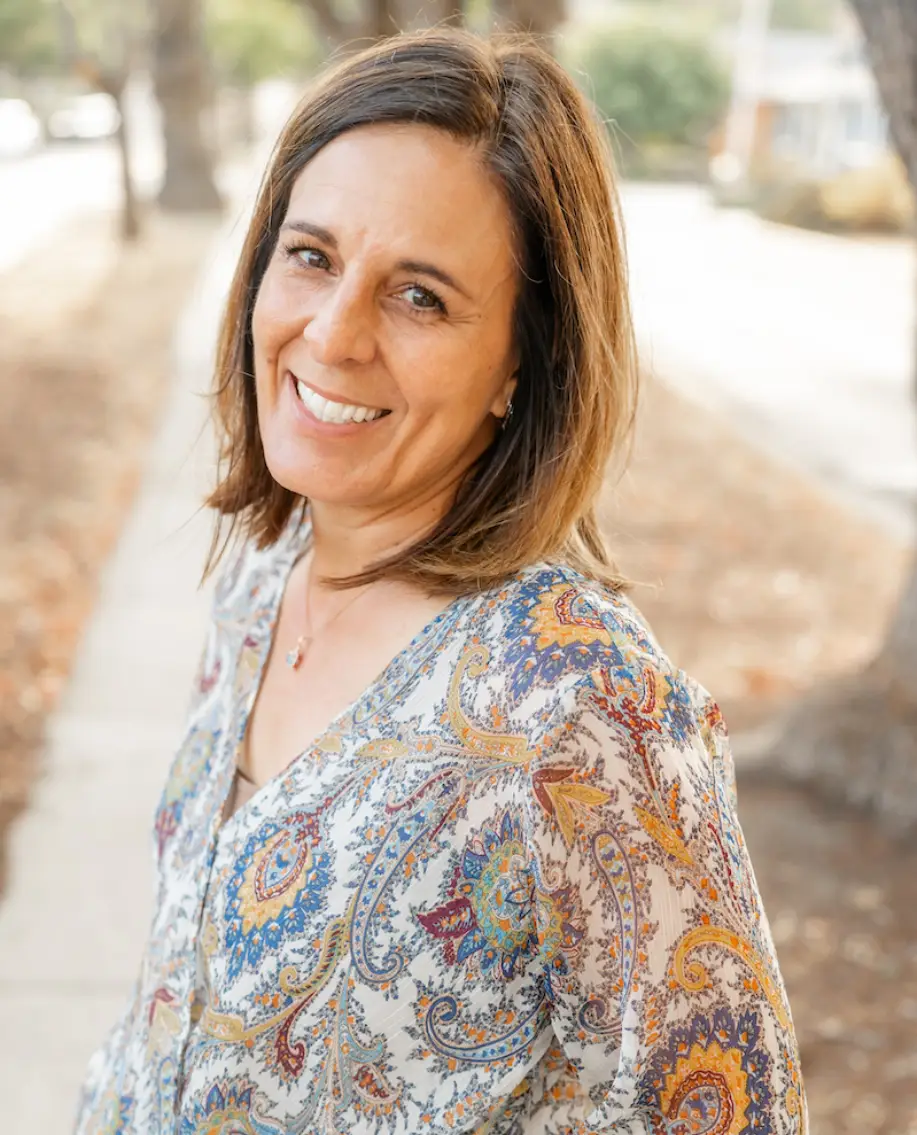 A woman stands outdoors wearing a colorful, patterned blouse, with trees and a sidewalk in the background.